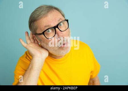 Listening to secrets with hand on ear. Caucasian man portrait on blue studio background. Beautiful male model in yellow shirt posing. Concept of human emotions, facial expression, sales, ad. Copyspace. Stock Photo