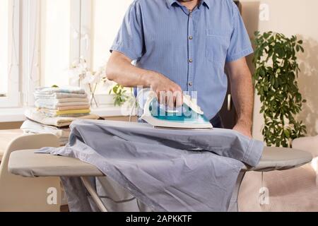 Man in the living room irons the sleeves of a shirt on an ironing stand on an ironing board. In the background, ironed and folded towels lie on the ta Stock Photo