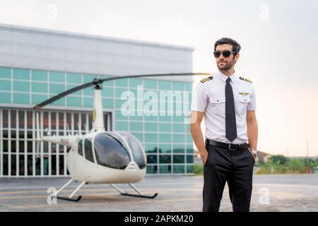 Portrait of handsome commercial pilot in captain white uniform standing and looking smart near small private helicopter on a landing point with privat Stock Photo
