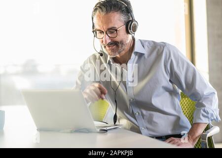Mature businessman wearing headphones using laptop at desk in office Stock Photo
