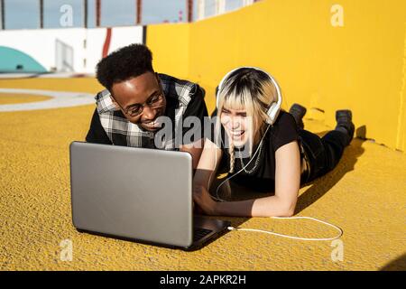 Laughing young couple using headphones and laptop outdoors having fun Stock Photo