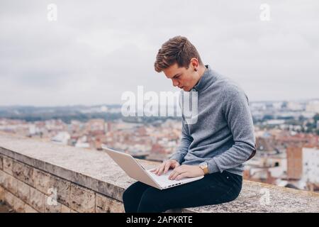 Blond young man sitting on a wall and typing on his laptop in Lleida Stock Photo