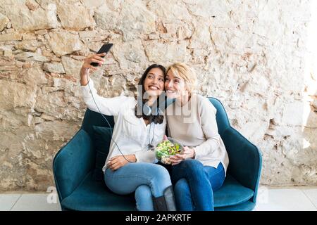 Two colleagues sitting on couch at lunchtime taking selfie with smartphone Stock Photo