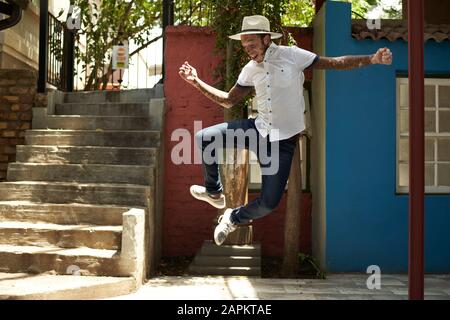 Portrait of young man with vitiligo wearing a hat, jumping in the street Stock Photo