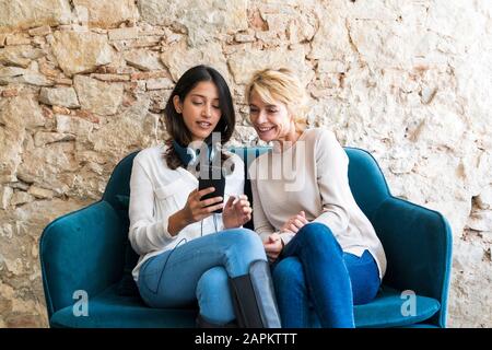 Portrait of two colleagues sitting on couch at looking at smartphone Stock Photo