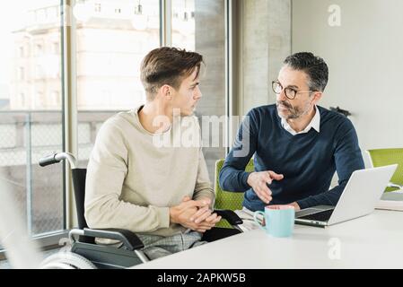 Mature businessman and young man in wheelchair talking at desk in office Stock Photo