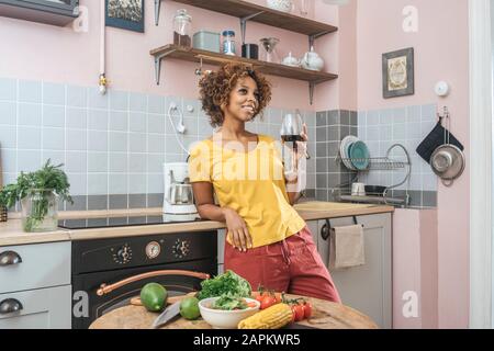 Smiling young woman drinking glass of red wine in kitchen Stock Photo