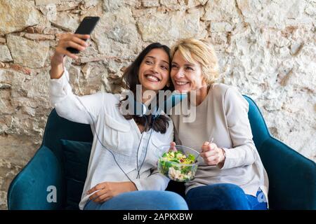 Two happy colleagues sitting on couch at lunchtime taking selfie with smartphone Stock Photo