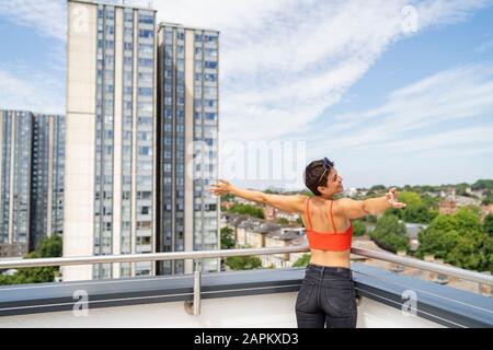 Back view of happy woman standing on roof terrace with arms outstretched Stock Photo