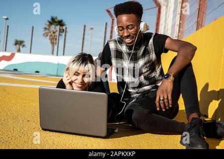 Portrait of smiling young couple using headphones and laptop outdoors Stock Photo