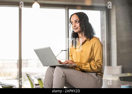 Young businesswoman sitting on desk in office using laptop Stock Photo
