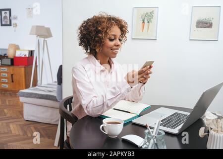 Young woman sitting at table at home using smartphone Stock Photo