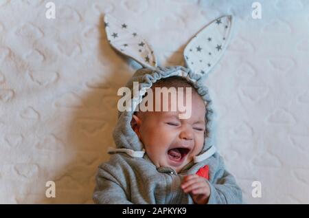Portrait of crying baby girl in a rabbit hoodie lying on white blanket Stock Photo