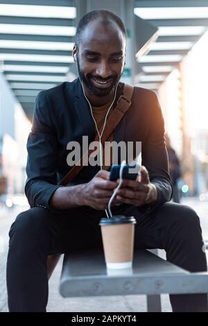 Smiling young businessman sitting at tram stop using earphones and smartphone Stock Photo