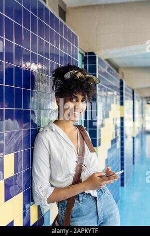 Portrait of smiling young woman with smartphone leaning against tiled wall Stock Photo