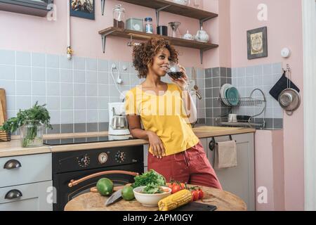 Smiling young woman drinking glass of red wine in kitchen Stock Photo