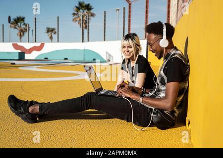 Smiling young couple using headphones and laptop outdoors Stock Photo