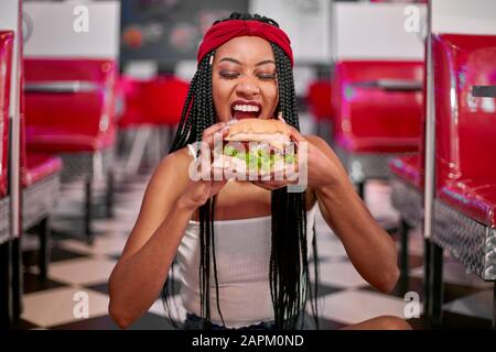 Young woman with braided hairstyle sitting on the floor and grabbing a big hamburger with her two hands Stock Photo