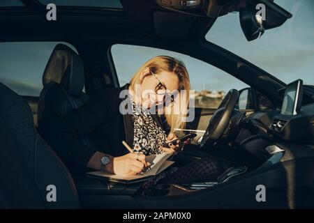 Young blond woman using smartphone in the car, writing in a notebook Stock Photo