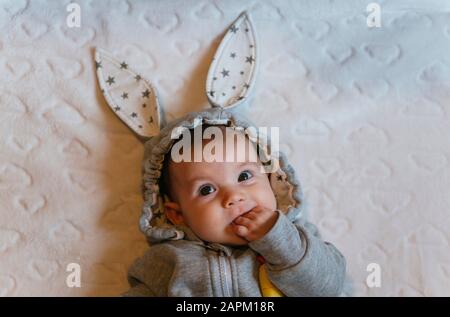 Portrait of baby girl in a rabbit hoodie with finger in mouth lying on white blanket Stock Photo