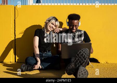 Portrait of laughing young couple sitting in front of yellow wall using headphones and laptop Stock Photo