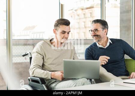 Mature businessman and young man in wheelchair using laptop in office Stock Photo
