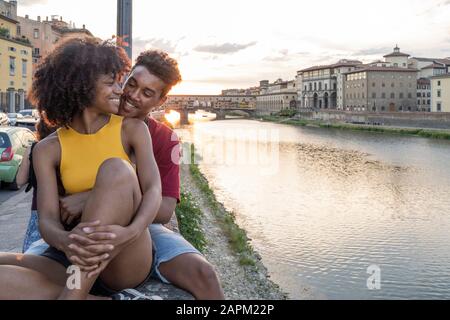 Affectionate young tourist couple sitting on a wall at river Arno at sunset, Florence, Italy Stock Photo
