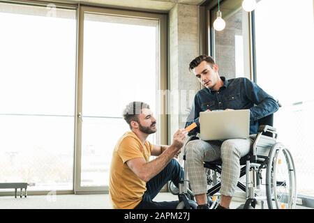 Young businessman in wheelchair and colleague using laptop in office Stock Photo