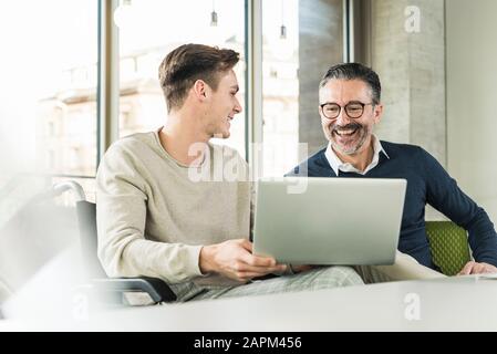 Happy mature businessman and young man in wheelchair using laptop in office Stock Photo
