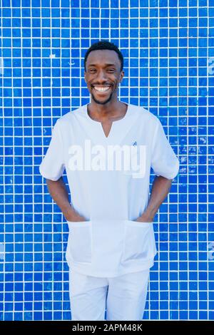 Portrait of laughing doctor standing against blue tiled wall with hands in his pockets Stock Photo