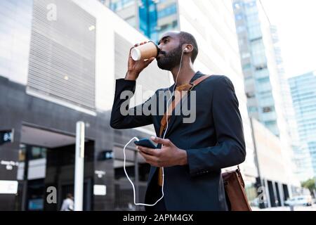 Young businessman with earphones and smartphone drinking coffee to go  outdoors Stock Photo