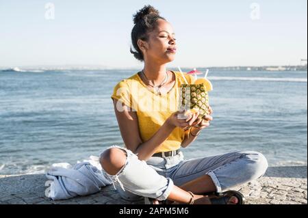 Young woman sitting at the waterfront enjoying a drink in a pineapple, Lisbon, Portugal Stock Photo