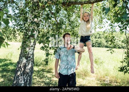Father checking the time with daughter hanging on a branch of a birch tree Stock Photo