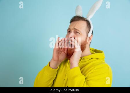 Calling, screaming. Caucasian man as an Easter bunny with bright casual clothes on blue studio background. Happy easter greetings. Concept of human emotions, facial expression, holidays. Copyspace. Stock Photo