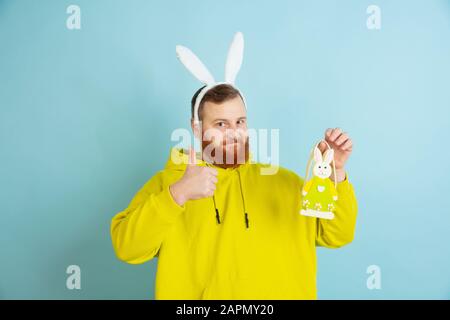 Pointing on decoration. Caucasian man as an Easter bunny with bright casual clothes on blue studio background. Happy easter greetings. Concept of human emotions, facial expression, holidays. Copyspace. Stock Photo