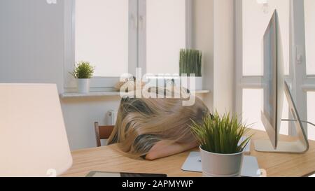 Tired blond female office worker in elegant suit relaxing lying on arms, on desk then getting up yawning and starting working on computer in light room with plants. Stock Photo