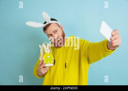 Caucasian man as an Easter bunny with bright casual clothes and decor taking selfie on blue studio background. Happy easter greetings. Concept of human emotions, facial expression, holidays. Copyspace. Stock Photo