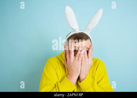 Scared, hiding face in hands. Caucasian man as an Easter bunny with bright casual clothes on blue background. Happy easter greetings. Concept of human emotions, facial expression, holidays. Copyspace. Stock Photo
