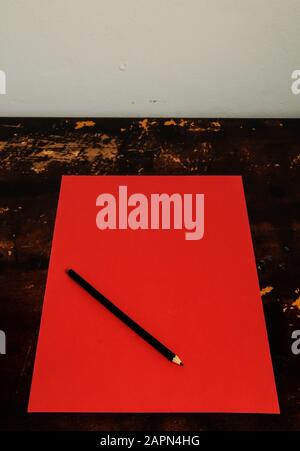 Overhead shot of a black pencil placed at the top of a red paper on a brown table Stock Photo