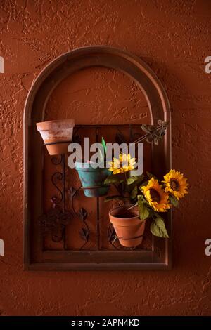 A wall-hung flower arrangement of three terra-cotta flower pots and three sunflowers with an arch frame, hangs on an outdoor wall that is painted red. Stock Photo