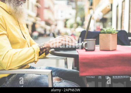Trendy businessman using laptop in bar cafeteria outdoor - Mature hipster male browsing online and drinking coffee - Technology and fashion lifestyle Stock Photo