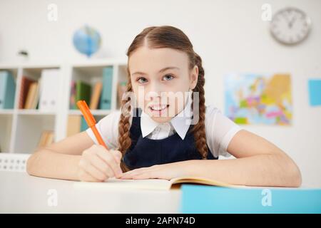 Portrait of schoolgirl sitting at desk and looking at camera while making notes in notebook during lesson at school Stock Photo