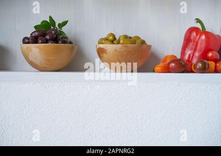 still life olives green brown and black in bowl with vegan red vegetables like tomatoes and paprika, white wall copy space vitamins Stock Photo