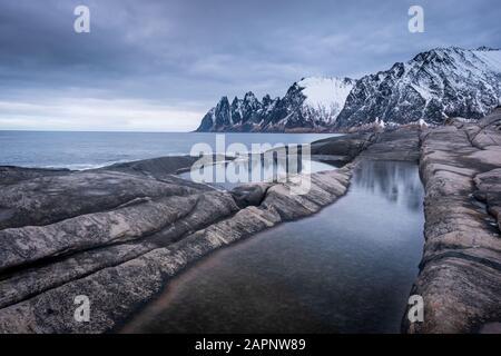 View over Ersfjord from colorful rocks and rockpools to snowy Oksen mountains on a dark cloudy day in Winter, Cape Tungeneset Senja Norway Stock Photo