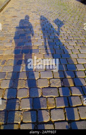 Dark, elongated shadows of man and woman on an old cobblestone pavement on a sunny day. Stones are colorful and reflect the sun rays.
