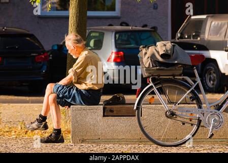 An old man sits on a bench, takes a break from cycling, old man enjoys the sun Stock Photo