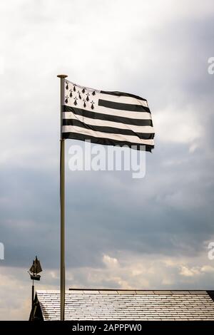 Flag of Brittany, named Gwenn ha Du in Breton, flying above a slate roof with a weather vane in the shape of a sailing ship against a stormy sky. Stock Photo