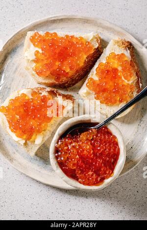 Red salmon caviar in bowl and on wheat bread, served on ceramic plate over grey spotted background. Flat lay, space Stock Photo