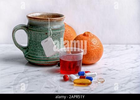 A green teacup, oranges, red cough syrup, and assorted pills are aligned on a white marble tabletop against a white background. Stock Photo