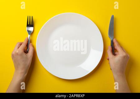 Female hands hold fork and knife on yellow background with plate, top view Stock Photo